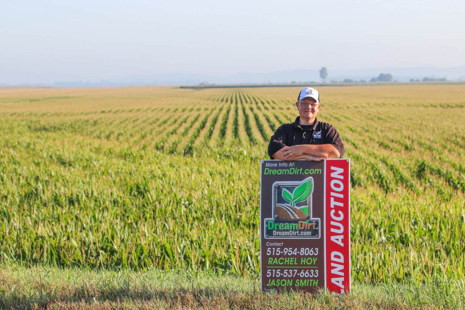 A DreamDirt auctioneer poses in a field with a Land Auction sign