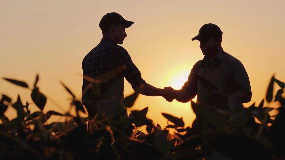 farmer and manager shake hands as the sun sets in a field.