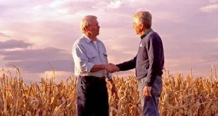 Two men shake hands in a corn field.